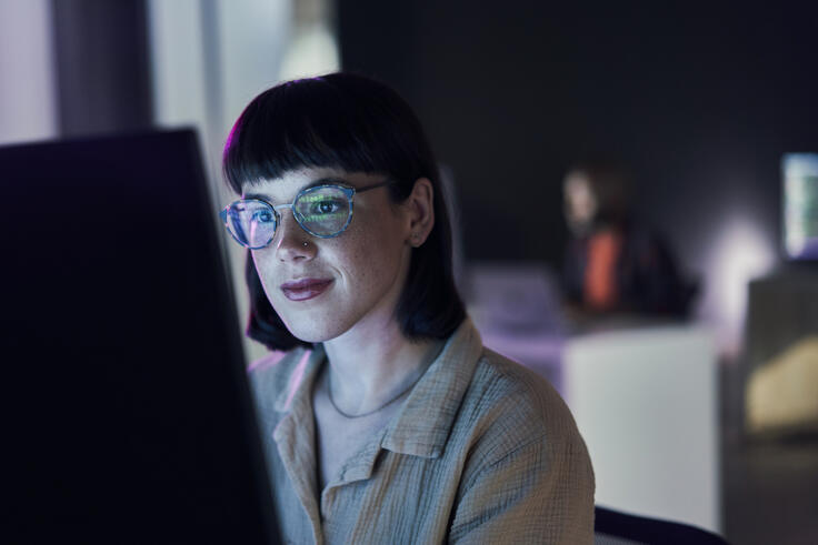 Young woman with glasses using a computer in a dimly lit office.