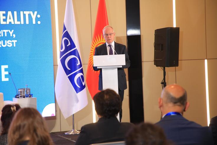 A man speaking at a podium at the OSCE RFoM's Central Asia Media Conference with a blue OSCE flag and a Kyrgyzstan flag beside him, audience in foreground.
