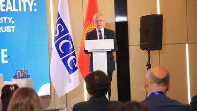 A man speaking at a podium at the OSCE RFoM's Central Asia Media Conference with a blue OSCE flag and a Kyrgyzstan flag beside him, audience in foreground.