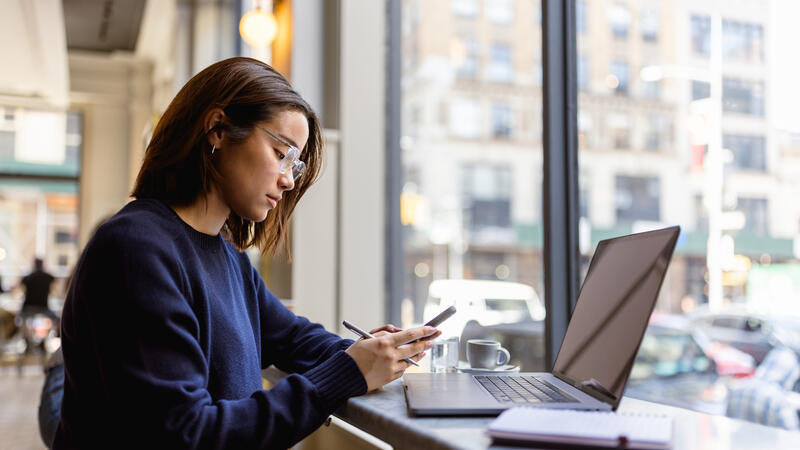 Woman working at a cafe with a laptop, smartphone, and notebook, focused and surrounded by large windows and city views.
